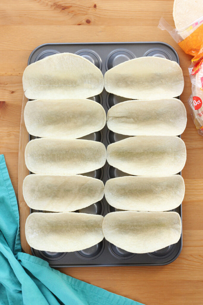 top down image showing raw tortillas placed in an upside down cupcake tin sitting on a wooden table with more tortillas and a teal napkin off to the side