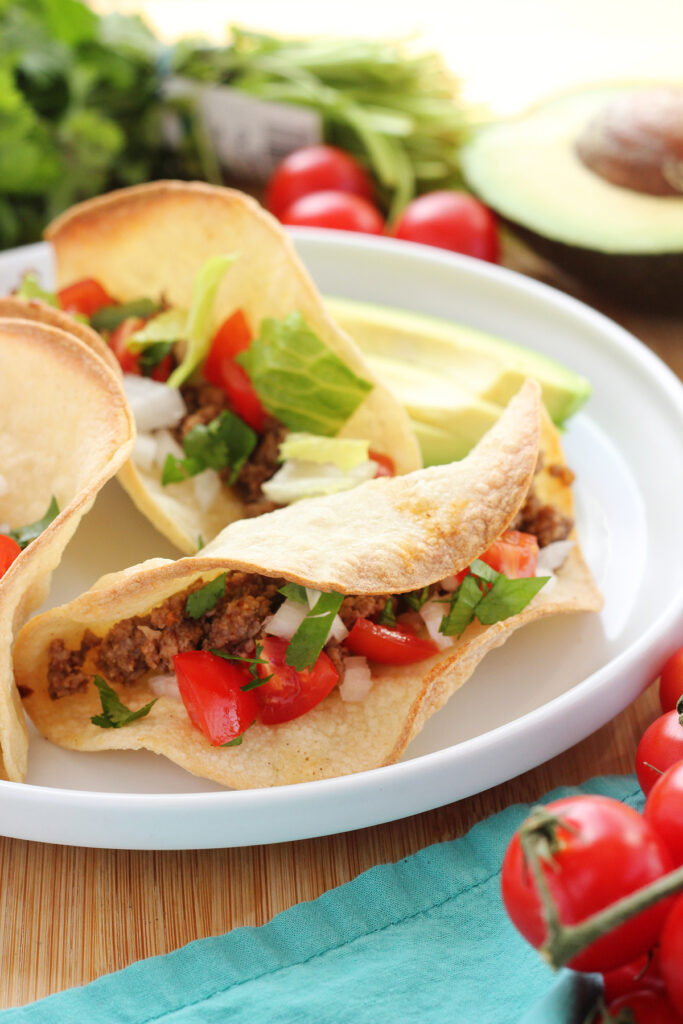 close up of three beef tacos filled with diced onion, tomato, and lettuce on a white plate with avocado slices, cherry tomatoes, and cilantro off in the background