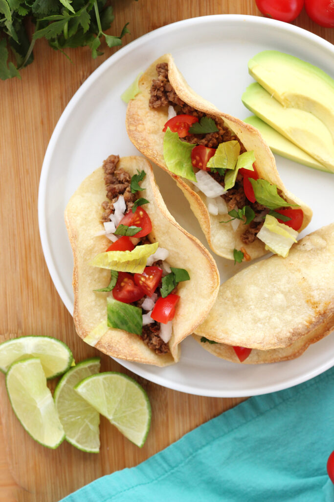 top down image showing a round white plate with three crispy tacos filled with beef, tomatoes, lettuce, and onion. There are avocado slices and lime wedges off to the side on a wooden cutting board