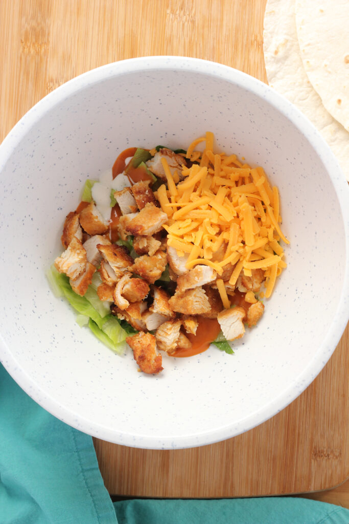 top down image showing a white mixing bowl with lettuce and drizzle of buffalo sauce and ranch with crispy chicken and shredded cheddar cheese