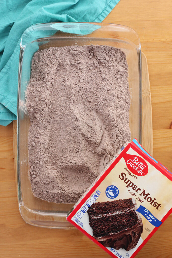 top down image of a 9x13 pan filled with dry cake mix sitting on a wooden table top. A box of betty crocker super moist chocolate cake sitting on the edge of the pan. 