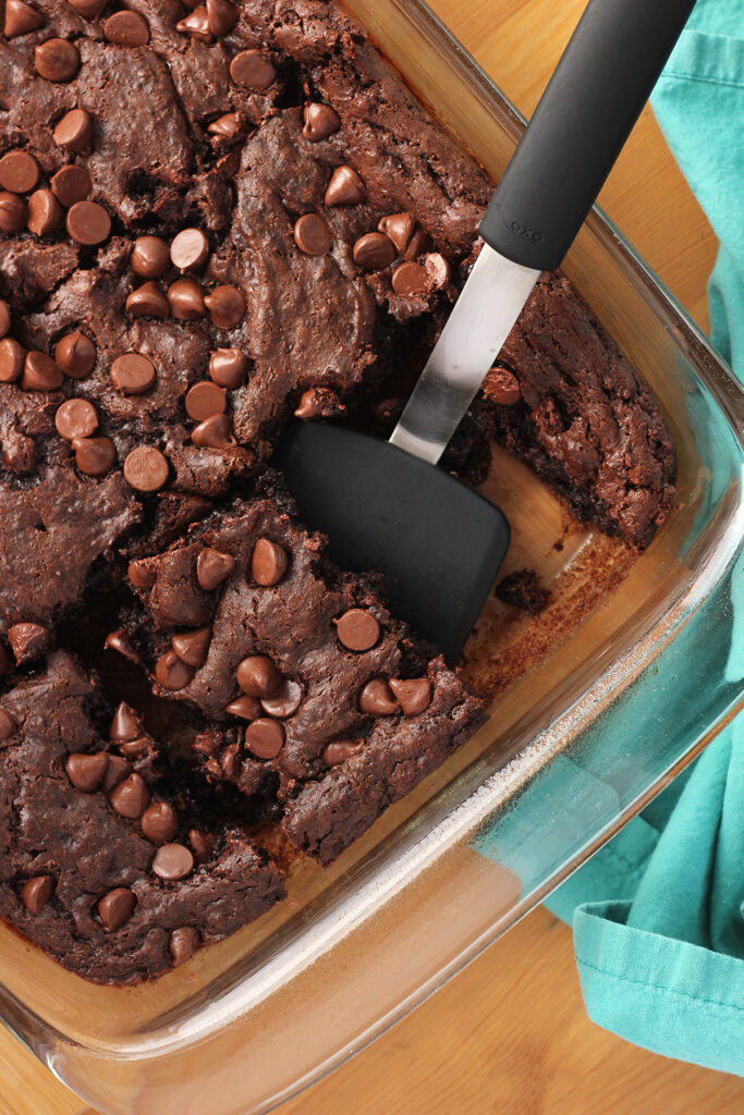 close up top down image showing a chocolate cake topped wtih chocolate chips inside a 9x13 pan with a slice being pulled out by a black spatula