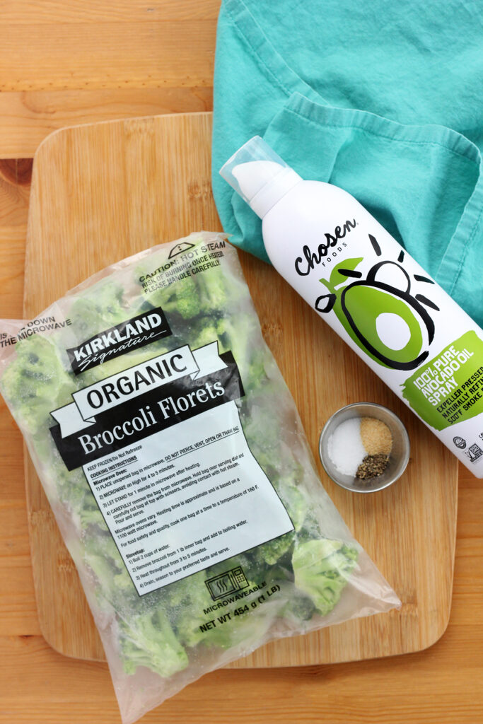 top down image showing a bag of Kirkland broccoli florets sitting on a wooden cutting board with a small silver bowl filled with seasonings and a can of avocado oil spray