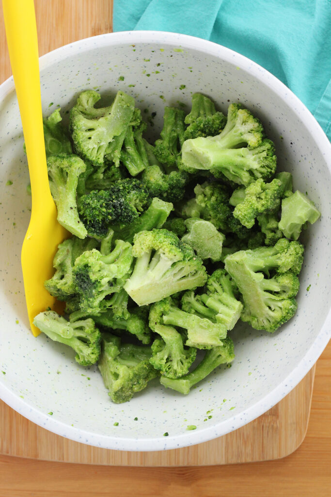 top down image showing a close up of a white mixing bowl filled with frozen broccoli and a yellow spatula