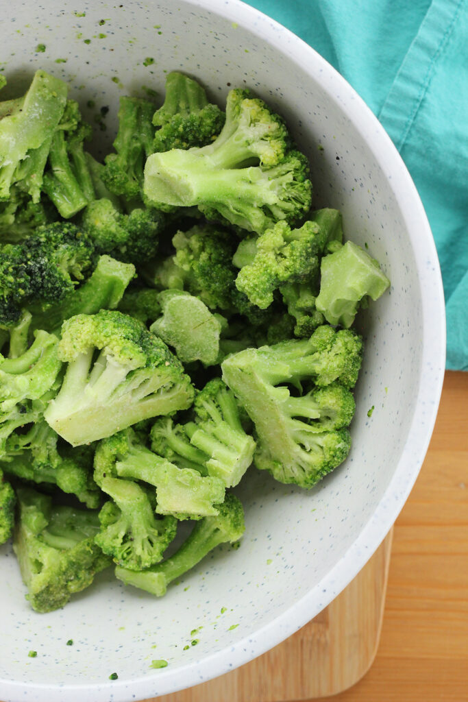 close up image showing a white mixing bowl that has frozen broccoli that has been coated in seasoning