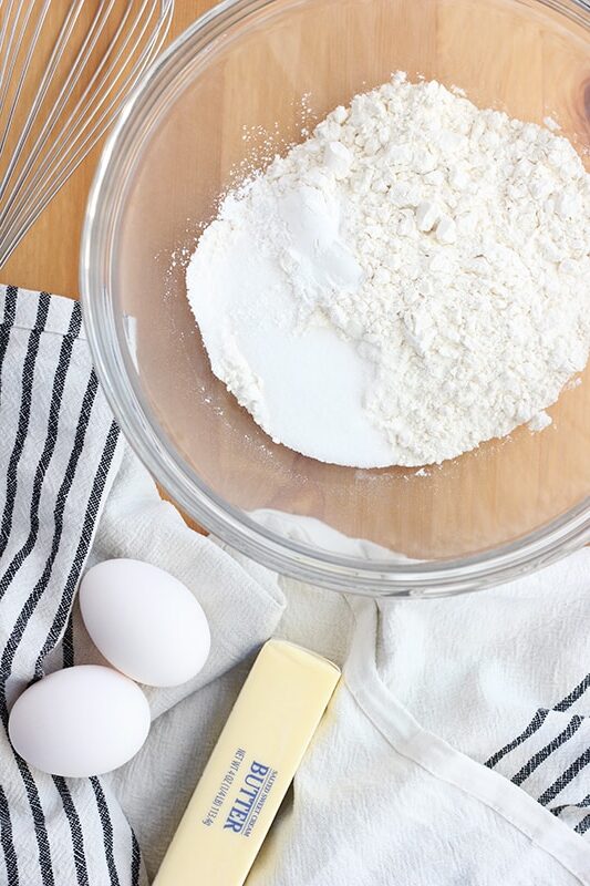 top down image showing a glass bowl filled with flour and sugar with a stick of butter and two eggs off to the side on a napkin with a whisk