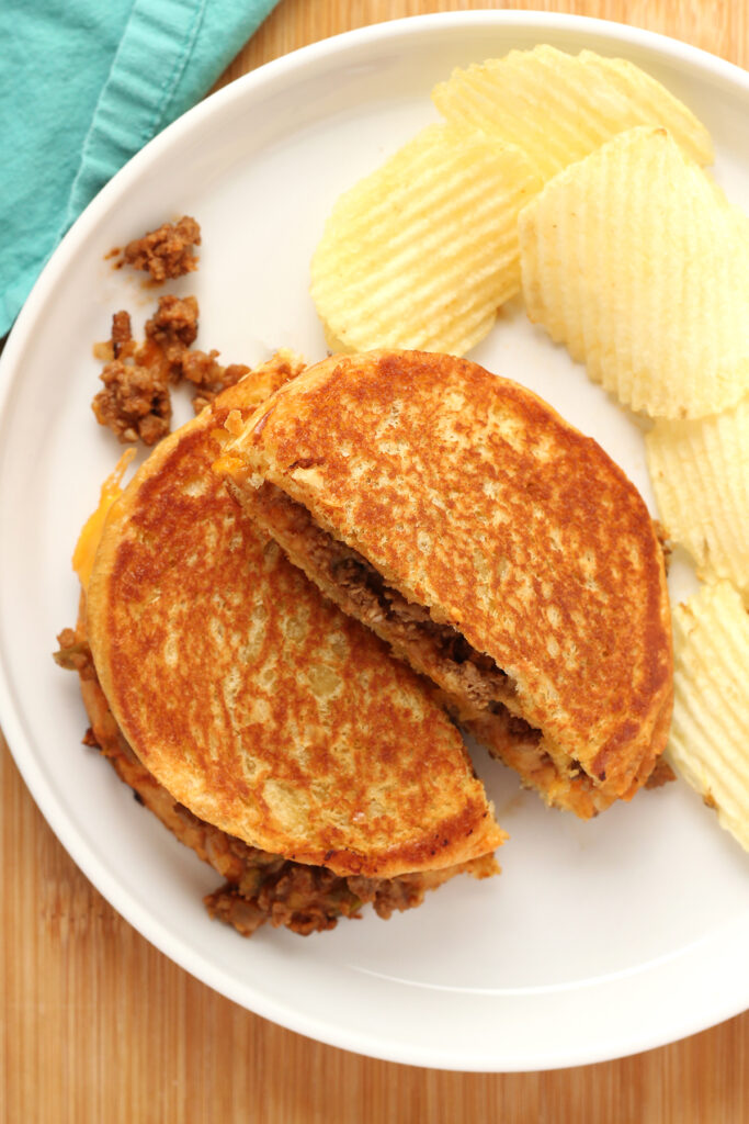 close up image showing a grilled sandwich with meat and cheese that has been cut in half on a white round plate with potato chips and a teal napkin off in the back