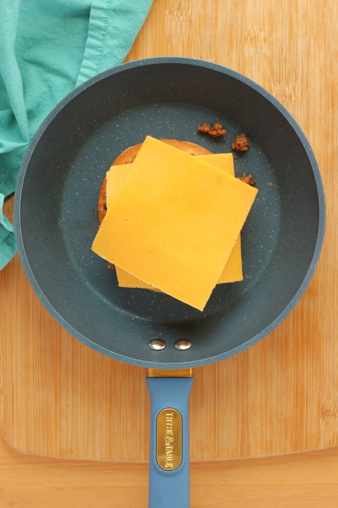 top down shot showing a blue skillet with an upside down bun topped with sloppy joe mix and cheese slices sitting on a wooden table top with a teal napkin off to the side. 