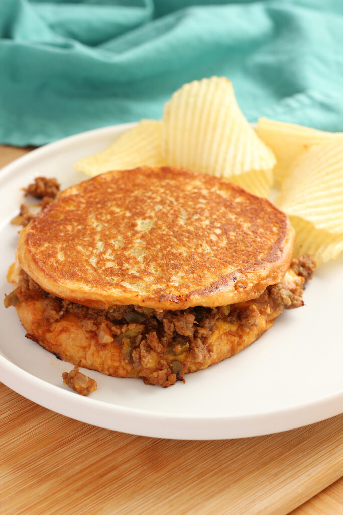 close up image showing a grilled sandwich with meat and cheese on a white round plate with potato chips and a teal napkin off in the back