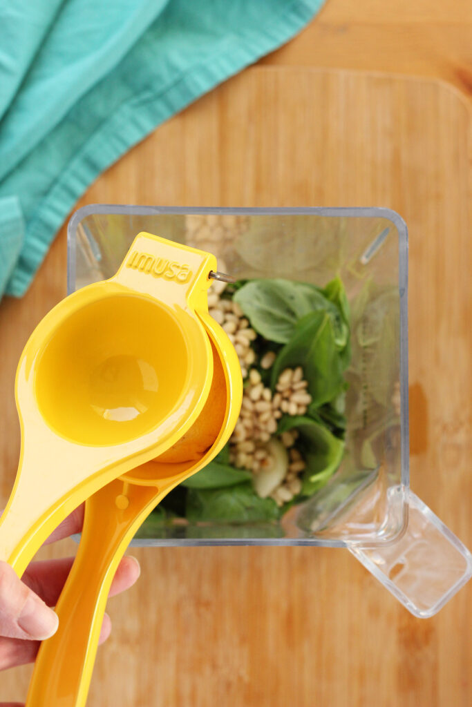 Top down image showing a blender that is filled with fresh basil and garlic cloves and pine nuts with a hand squeezing a yellow juicer filled with lemon