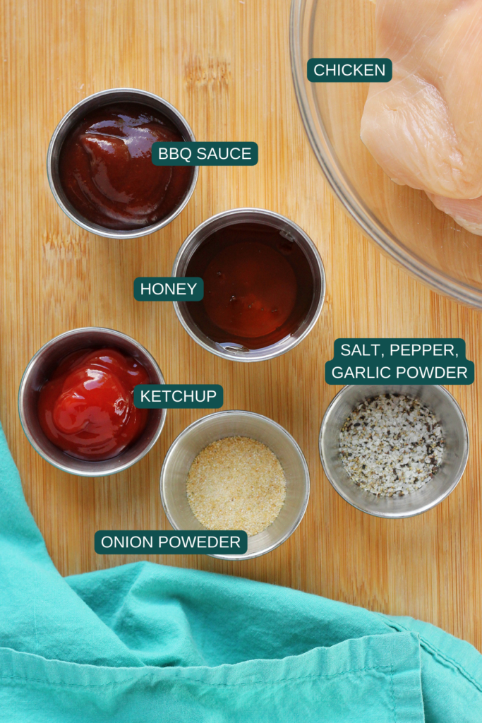 Top down image showing a glass bowl with chicken, and small ramekins filled with bbq sauce, honey, ketchup, salt, pepper, garlic powder, and onion powder on a wooden cutting board with a teal napkin