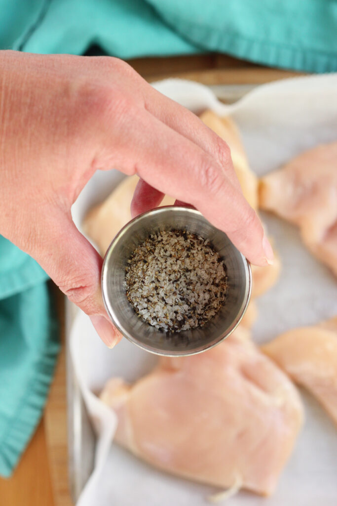 hand holding a small silver bowl of spices over raw chicken on a parchment lined tray