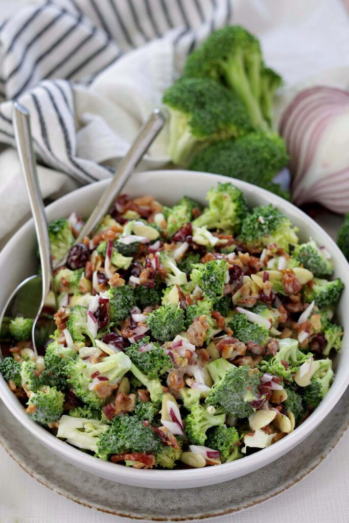 close up image showing a white bowl filled with broccoli that has been tossed in a dressing with dried cranberries, bacon pieces, and and almonds