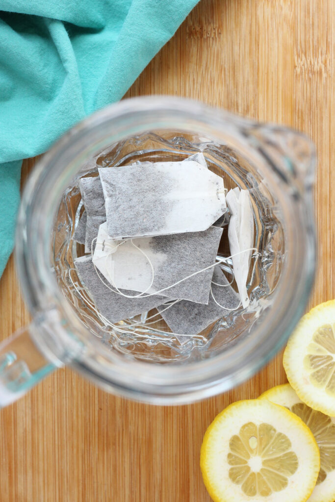 top down image showing a glass pitcher filled with tea bags sitting on a wooden cutting board with lemon slices and a teal napkin on the side