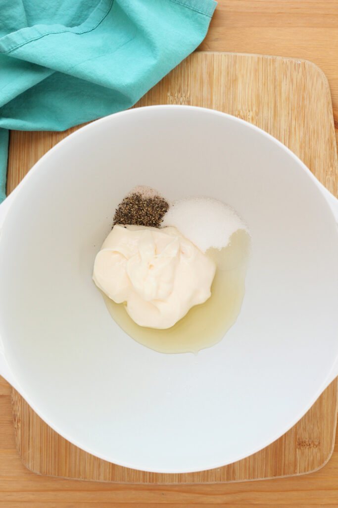 top down image showing a white mixing bowl on a wooden table top filled with mayo, salt and pepper, sugar, liquid