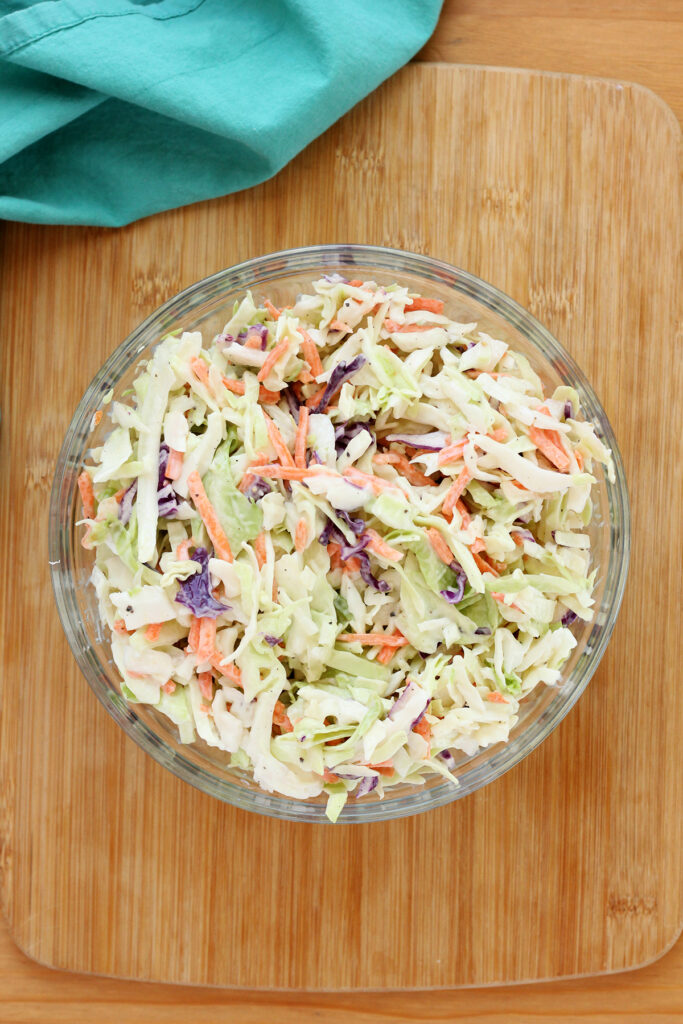 close up image of a glass mixing bowl filled with a slaw sitting on a wooden cutting board with a teal napkin off in the back
