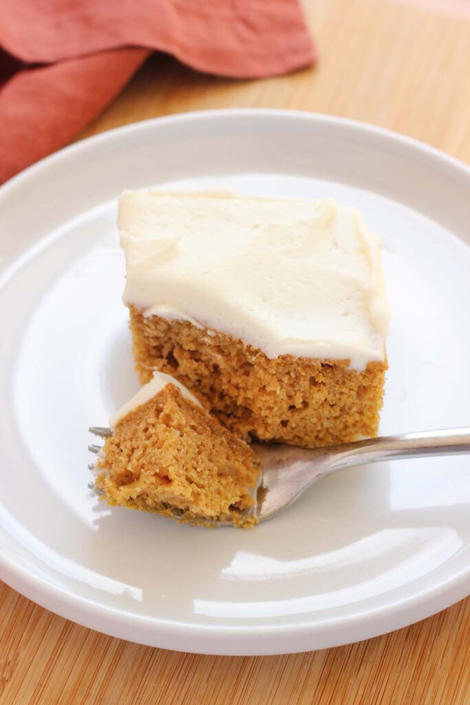 close up image of a piece of cake sitting on a white round plate with one bite on a fork. The cake is topped with a white frosting and a rust red napkin is off to the side