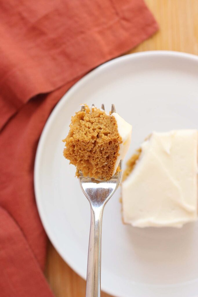 top down image showing a bite of pumpkin cake on a fork hovering over the top of a piece of cake on a white plate with a rust napkin off to the side