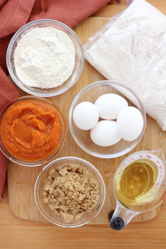 top down image showing a wooden table top with ingredients for a cake; cake mix, bowls filled with flour, eggs, pumpkin and brown sugar, and a cup of oil sitting with a rust colored napkin off to the side