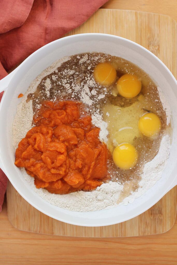 top down image showing a white mixing bowl filled with a dry cake mix, oil, eggs, and pumpkin that is not mixed together