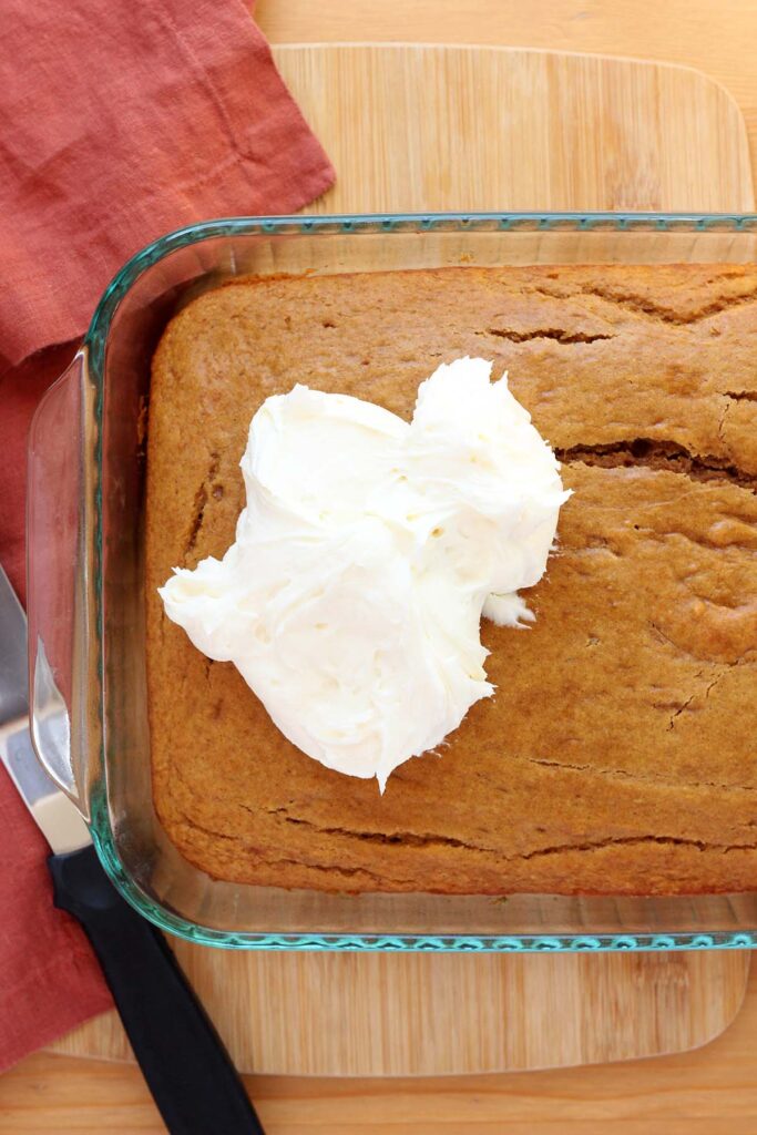 close up image showing a baked cake inside a glass pan with dollops of white frosting on top. The cake is sitting on a wooden table top with a rust colored napkin and a metal spatula sitting off to the side