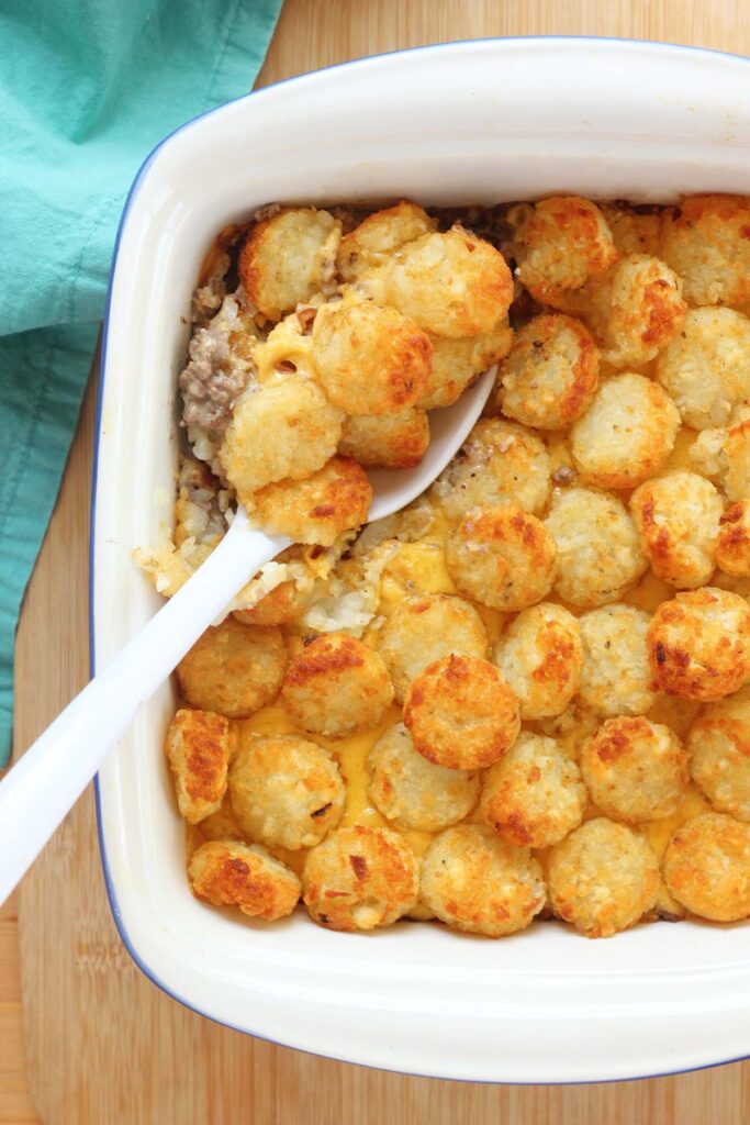 Top down image showing a white spoon scooping out a casserole topped with tater tots from a white baking dish