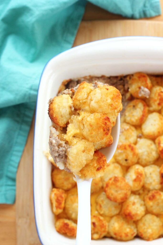 Top down image showing a white spoon scooping out a casserole topped with tater tots from a white baking dish