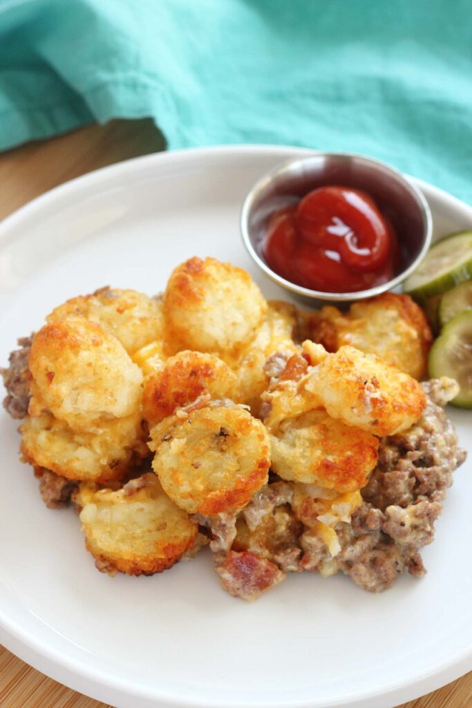 Close up image of a white round plate topped with a tater tot casserole with a side of bacon and ketchup. A teal napkin off in the background