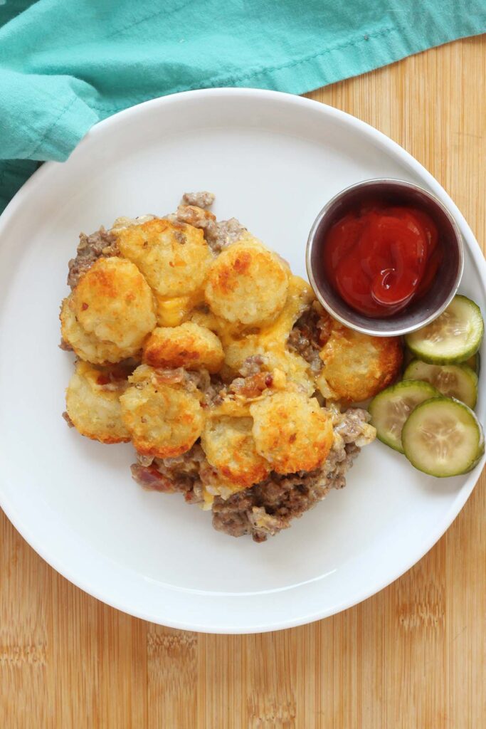 Close up image of a white round plate topped with a tater tot casserole with a side of bacon and ketchup. A teal napkin off in the background