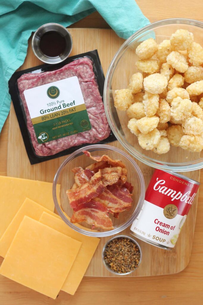 top down shot showing ingredients sitting on a wooden table top. There is a container of ground beef, small dish with a brown liquid, bowl of tater tots, bowl of bacon, can of cream of onion soup, container of seasoning, and four cheese slices