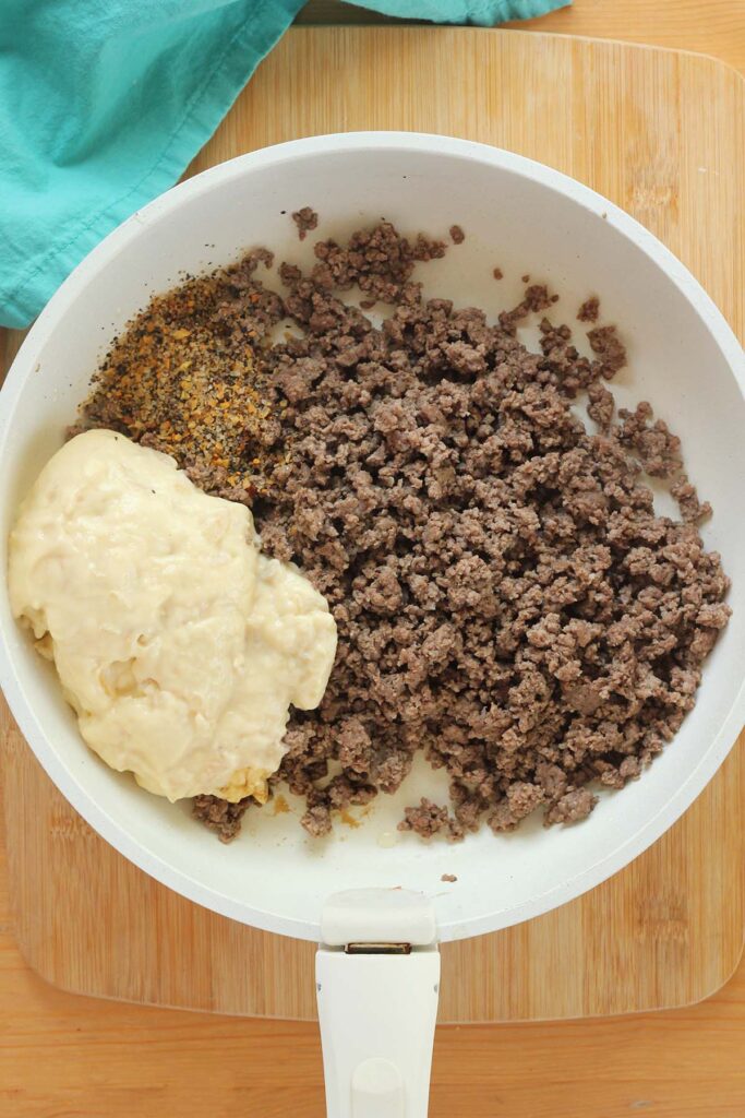 top down image showing a white pan filled with hamburger, spices, and cream soup on a wooden table top with a teal napkin