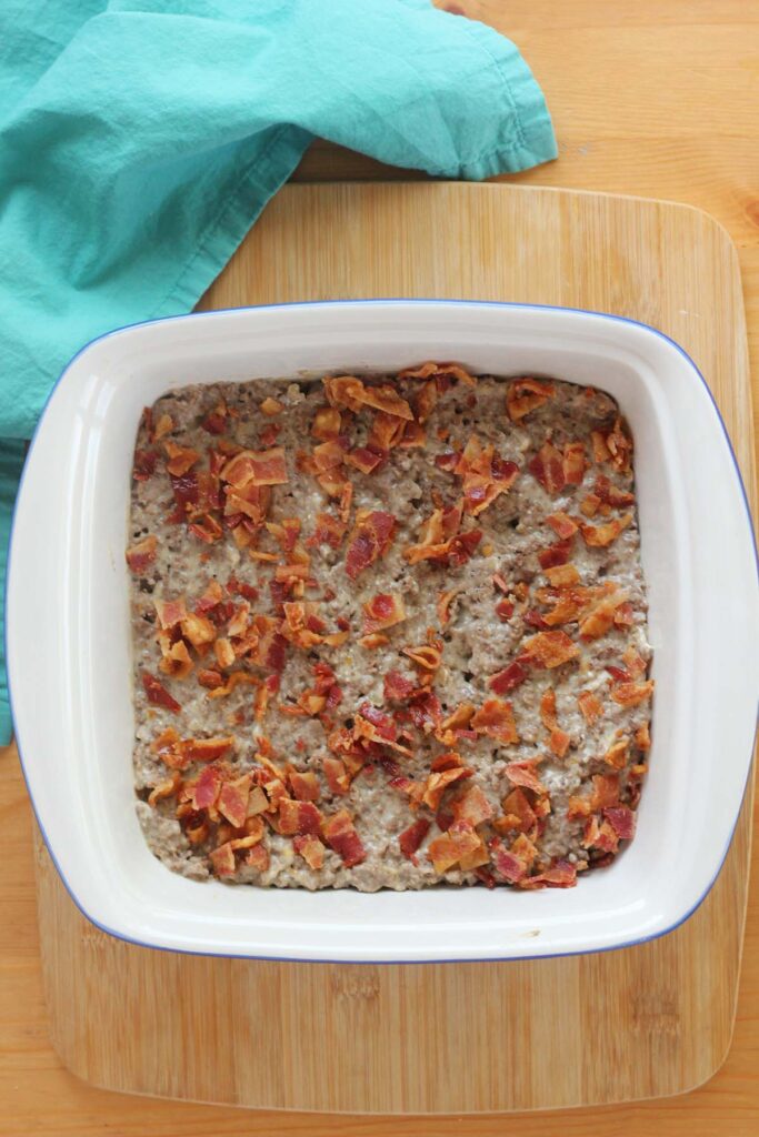 top down image showing a white baking dish filled with hamburger and bacon pieces sitting on a wooden table with a teal napkin