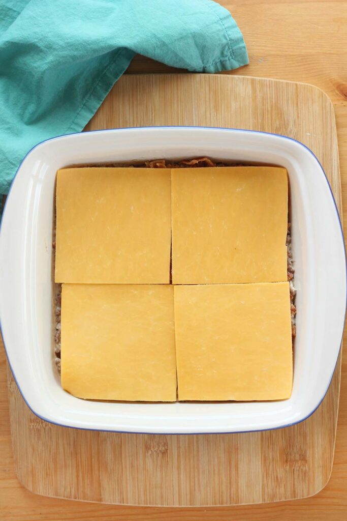 top down image showing a white baking dish filled with hamburger and cheese slices sitting on a wooden table with a teal napkin