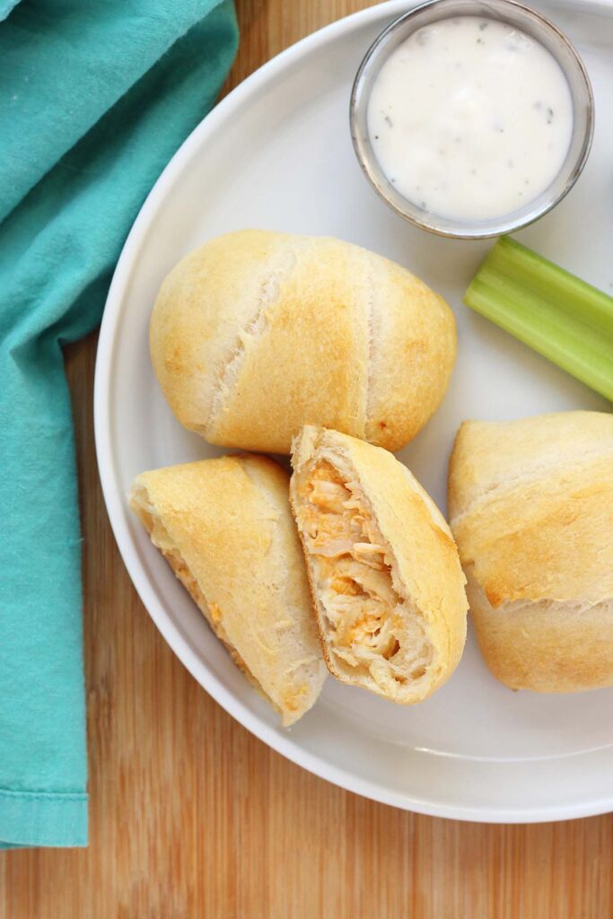 close up image showing three crescent pockets, one cut in half and filled with chicken and cheese, sitting on a white round plate with three celery sticks and a small dish of ranch dressing. The plate is sitting on a wooden table top with a teal napkin off in the back