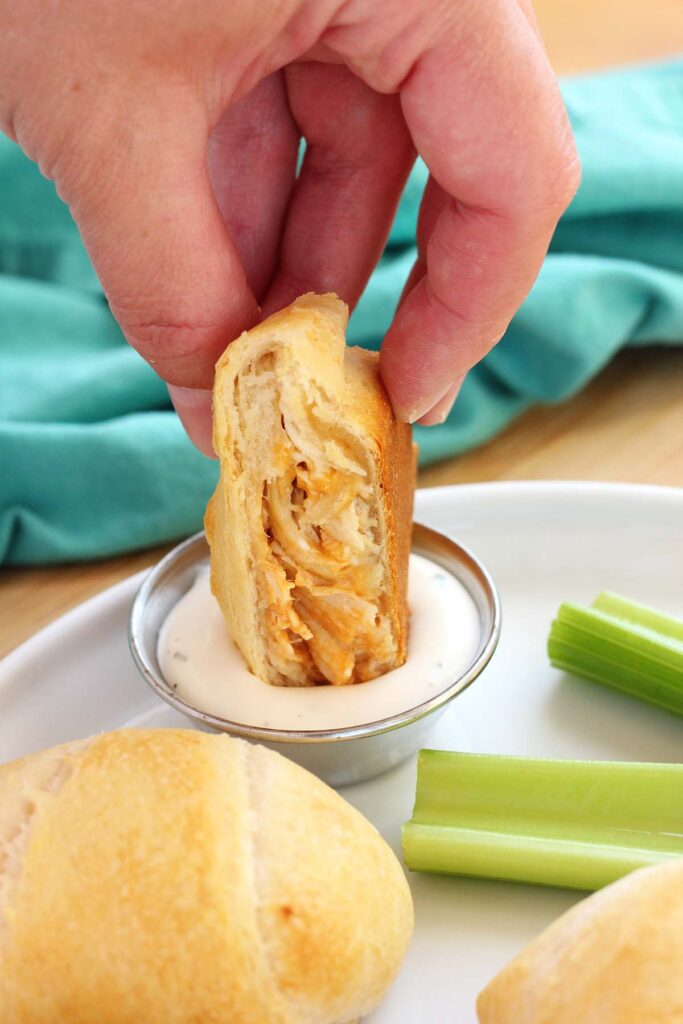 close up image showing three crescent pockets, one cut in half and filled with chicken and cheese, sitting on a white round plate with three celery sticks and a small dish of ranch dressing. The plate is sitting on a wooden table top with a teal napkin off in the back