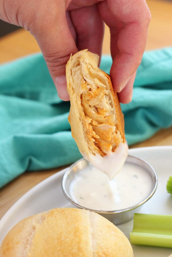 close up image showing three crescent pockets, one cut in half and filled with chicken and cheese, sitting on a white round plate with three celery sticks and a small dish of ranch dressing. The plate is sitting on a wooden table top with a teal napkin off in the back