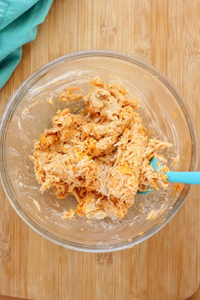 top down image showing a glass bowl with shredded chicken, buffalo sauce, shredded cheddar, and cream cheese that has been mixed with a blue spatula