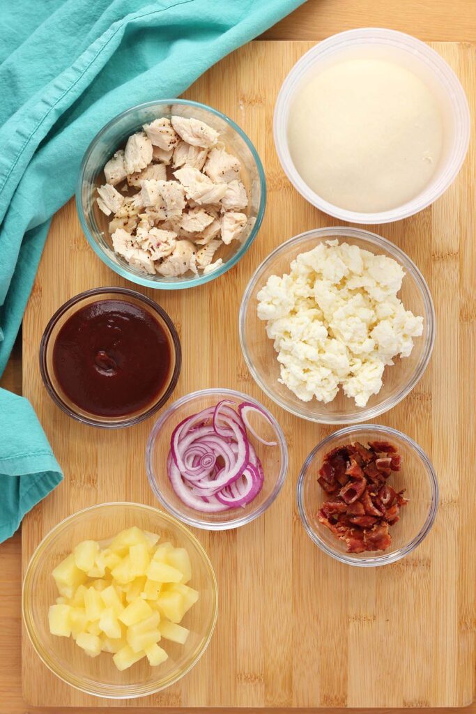 Top down image showing ingredients for a chicken pizza, including pizza dough, chopped chicken, bbq sauce, mozzarella cheese, sliced red onoin, bacon, and pineapple