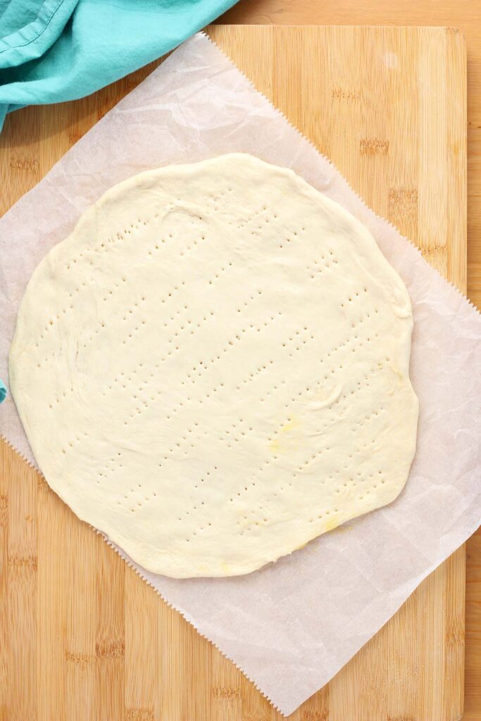 Top down image showing a pizza dough rolled out onto a piece of parchment paper sitting on a wooden counter with a teal napkin off to the side. 