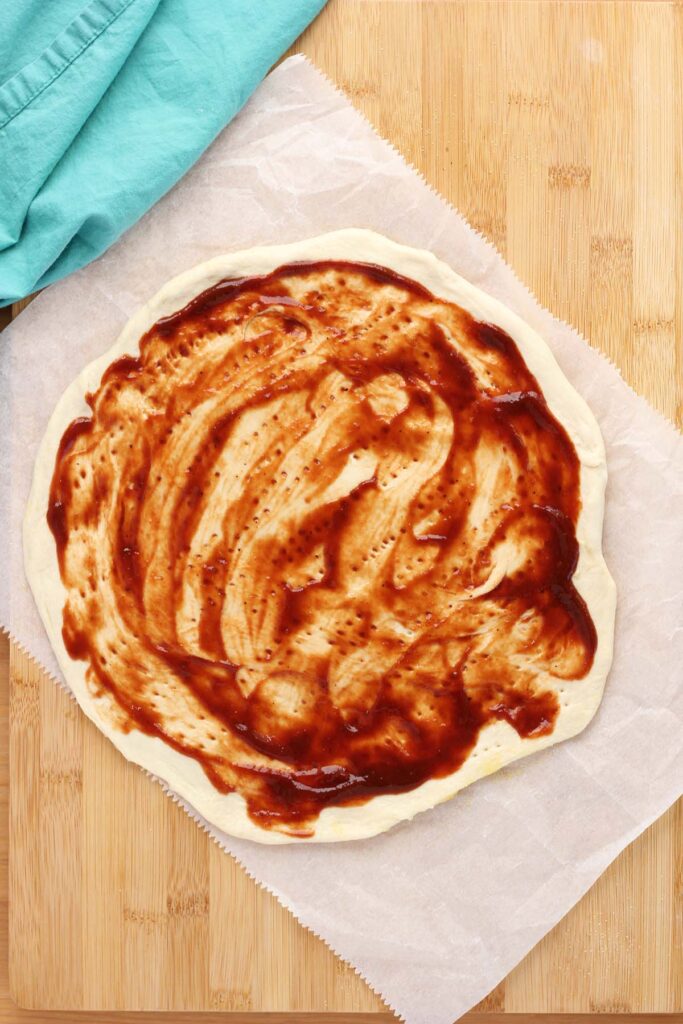 Top down image showing a pizza dough rolled out onto a piece of parchment paper with bbq sauce sitting on a wooden counter with a teal napkin off to the side. 