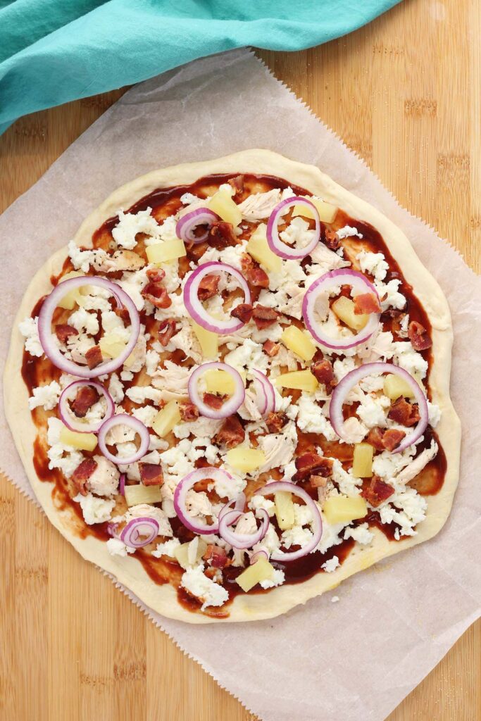 unbaked pizza sitting on a wooden table top covered in toppings ready to bake. There is cheese, chicken, pineapple, bacon, and red onion with a bbq sauce