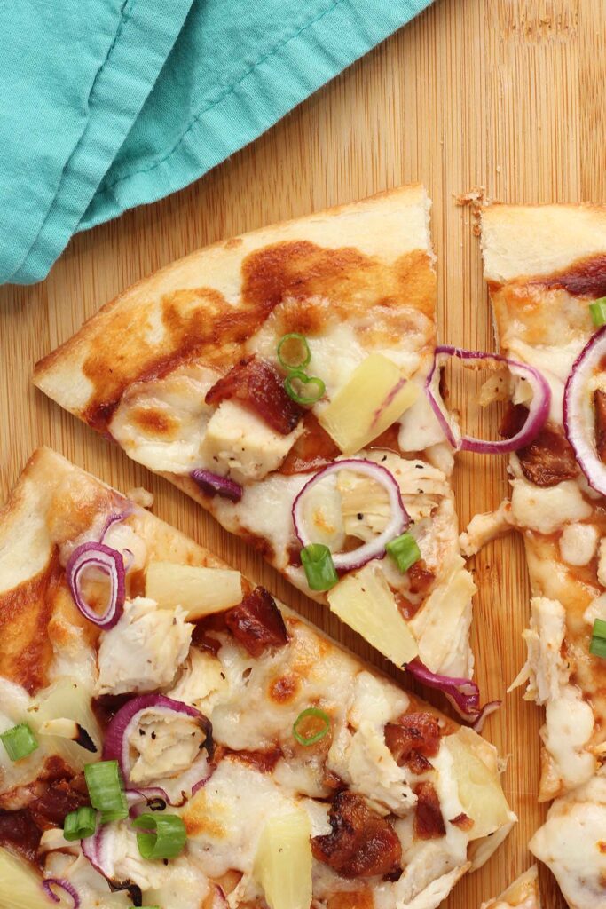 Top down image showing a baked pizza that is topped with cheese, chicken, pineapple ,red onion slices, and bacon. The pizza is sitting on a wooden table top with red onion slices and green onions off the side with a teal napkin