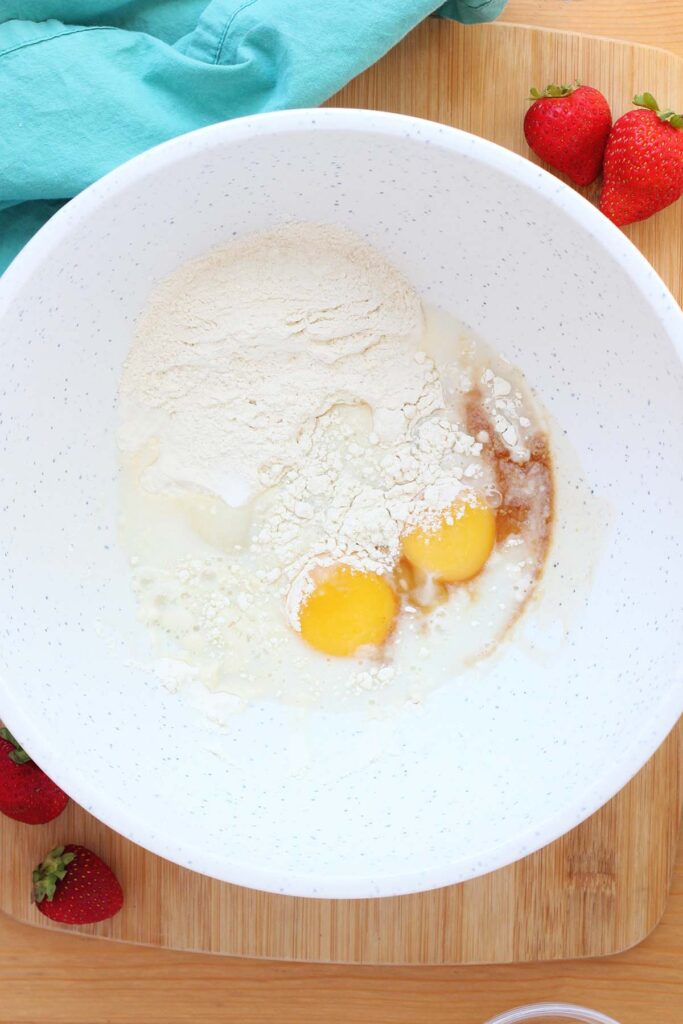 Top down image showing a white mixing bowl with flour, eggs, milk and vanilla not mixed together sitting on a wooden table top with strawberries and a teal napkin