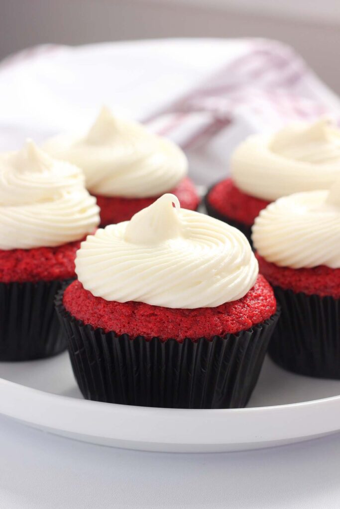 image showing a white round plate on a white table top filled with red cupcakes in black liners with white frosting