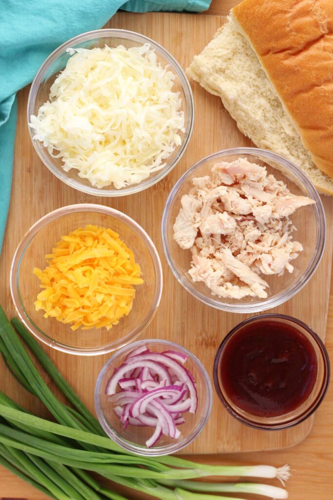Top down image showing a wooden cutting board with a sliced french bread, glass bowls each filled with either mozzarella cheese, chicken, cheddar, bbq sauce, red onion. Green onion and a teal napkin are off to the side