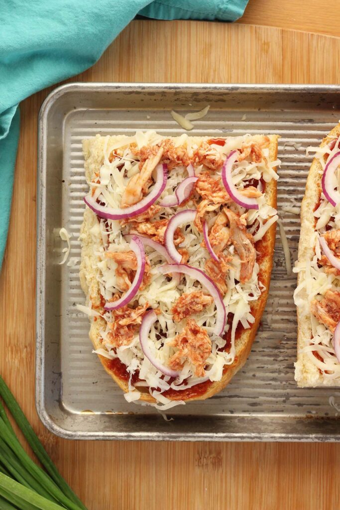 Top down image showing 1/2 a loaf of french bread sliced in half with bbq sauce spread across topped with cheese and chicken with red onion