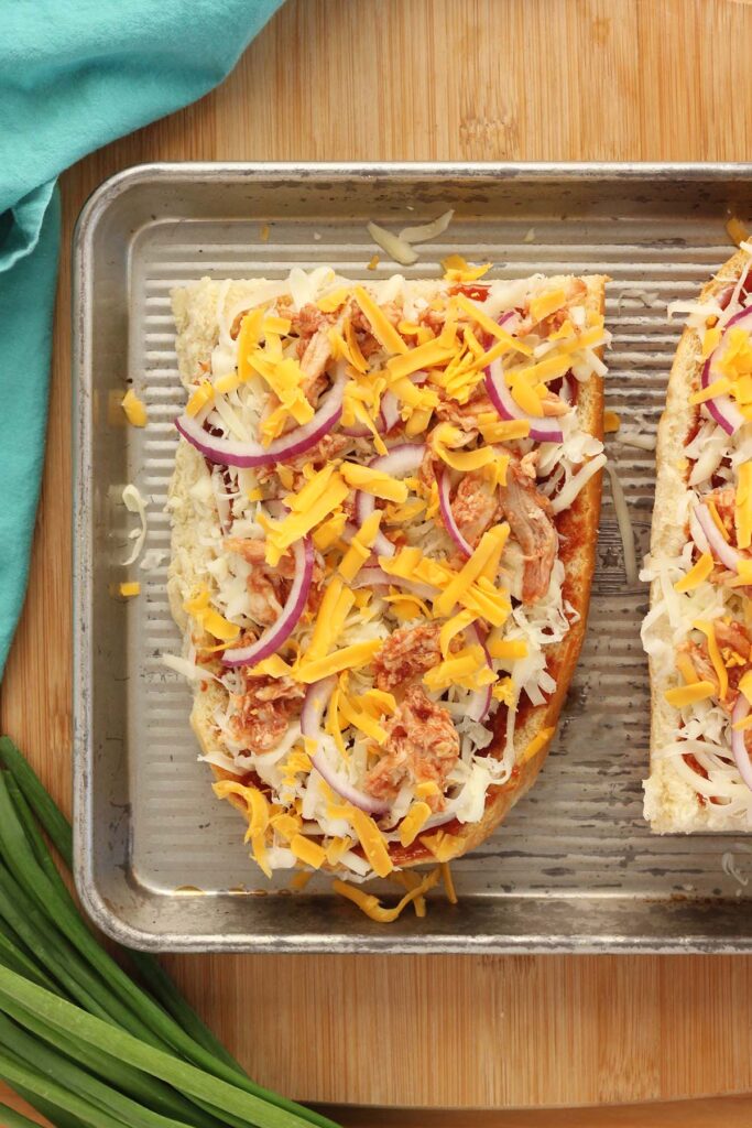 Top down image showing 1/2 a loaf of french bread sliced in half with bbq sauce spread across topped with cheese and chicken with red onion and a sprinkle of cheese