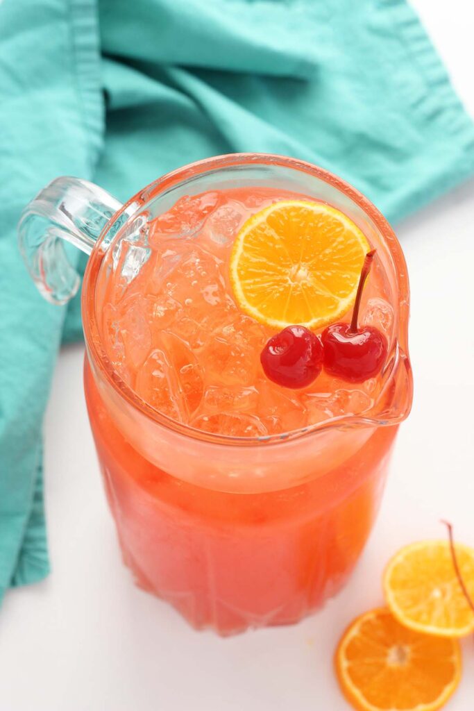 close up of a pitcher with a redish orange punch inside topped with two cherries and an orange slice