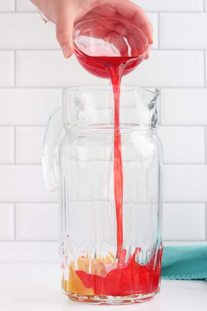 grenadine being poured into a glass pitcher with pineapple concentrate