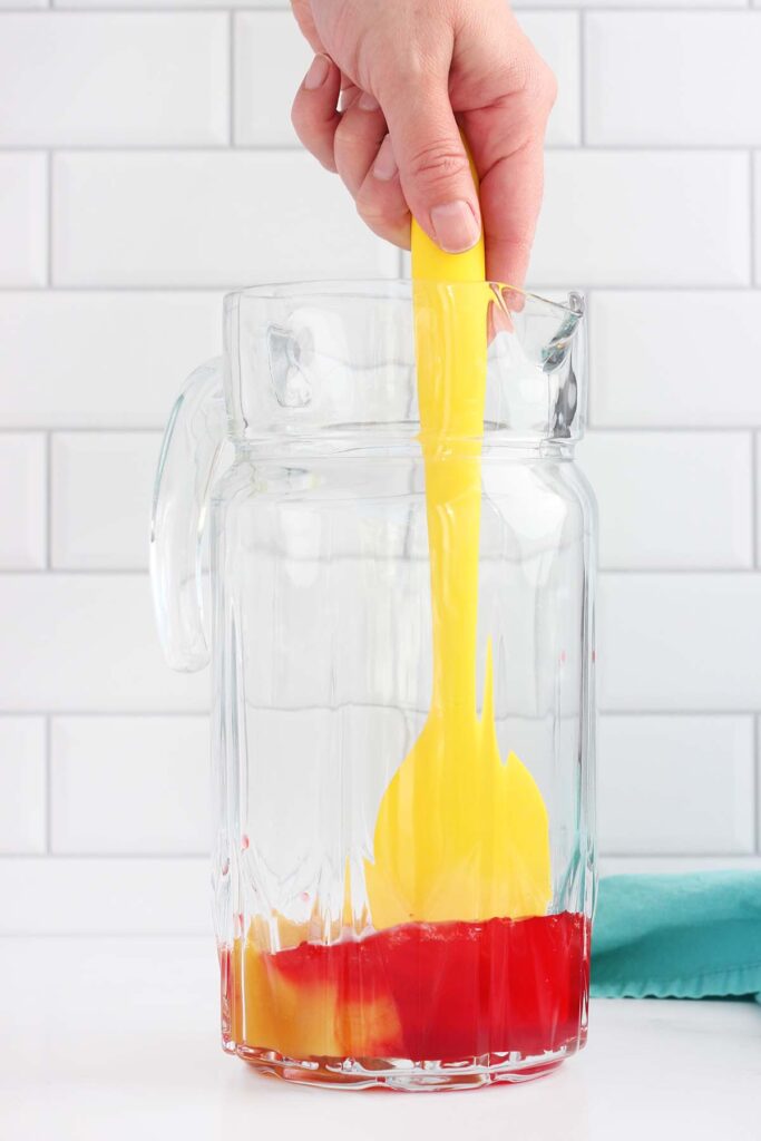 grenadine being mixed into a glass pitcher with pineapple concentrate using a yellow spatula